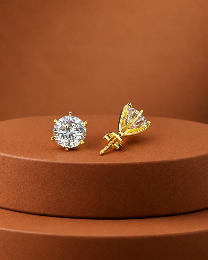 A pair of gold stud earrings displayed on a round brown pedestal. One earring is shown front-facing as a round brilliant-cut clear gemstone in a gold setting, and the other is shown from the side/back, revealing a prong design and screw-back post.