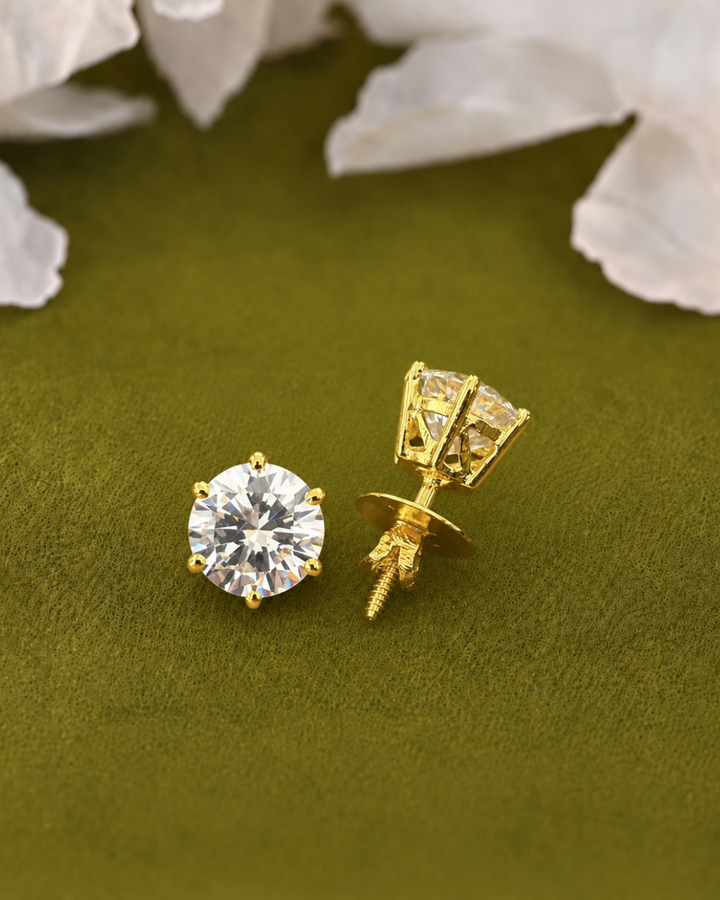 A pair of gold stud earrings, one showing a round clear gemstone in a prong setting and the other showing the back post and screw, displayed on an olive green textured surface with white flower petals in the background.