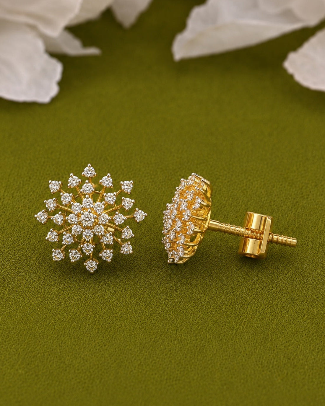 A pair of gold stud earrings with clear pavé-set stones, shown from both front and side angles on a textured olive green background with white flower petals.