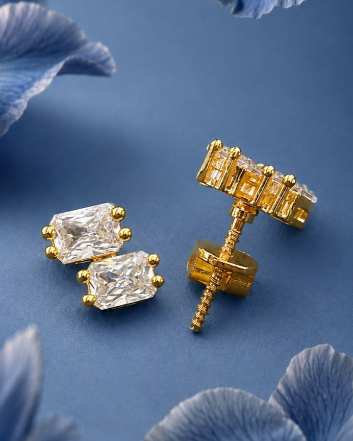 A pair of gold stud earrings with clear rectangular gemstones, shown with one earring front-facing and the other turned to show the post and backing, on a blue background with soft blue flower petals.