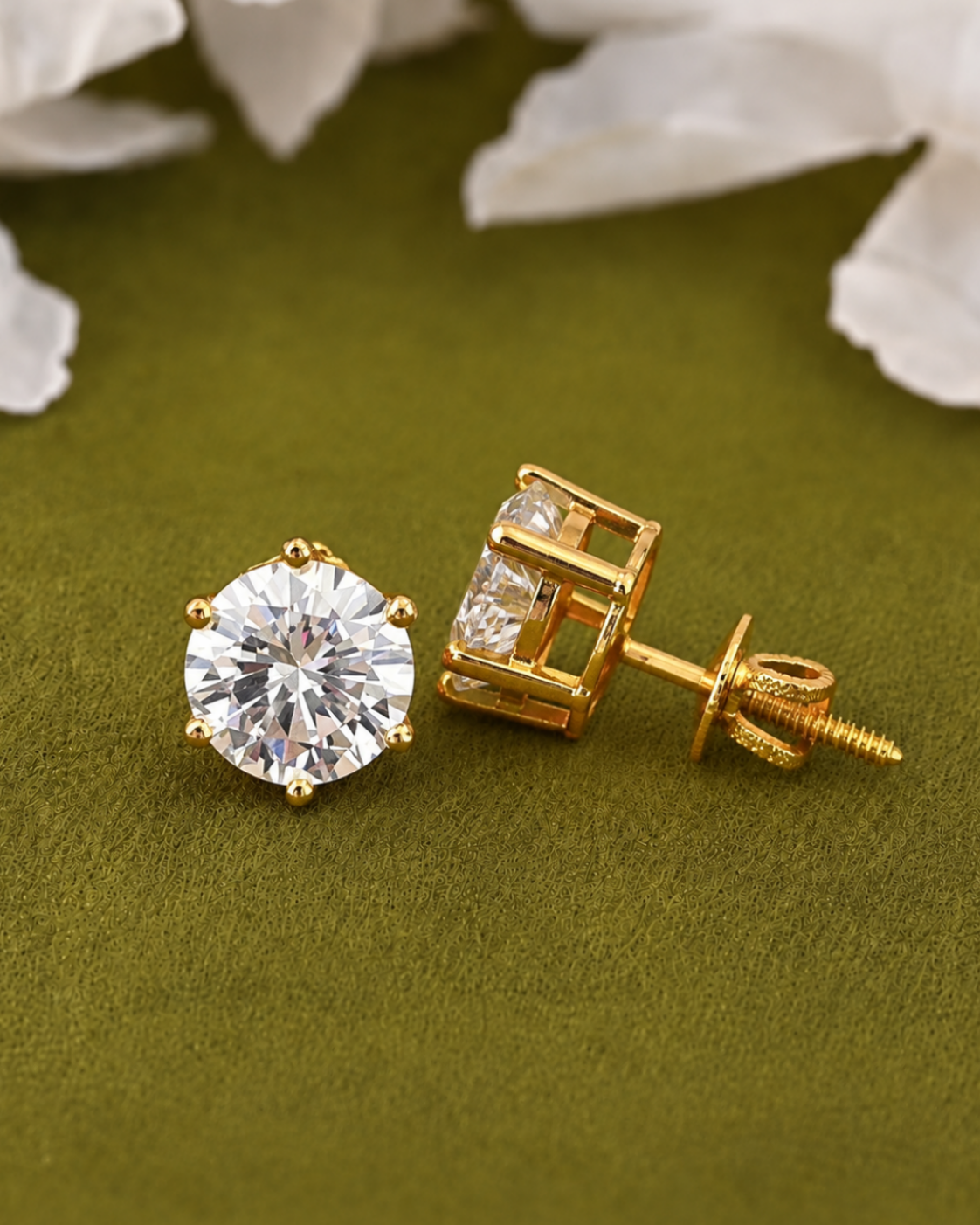 A pair of gold stud earrings, with one earring showing a round clear gemstone and the other showing the back with a screw post, displayed on an olive green textured surface with white flower petals in the background.