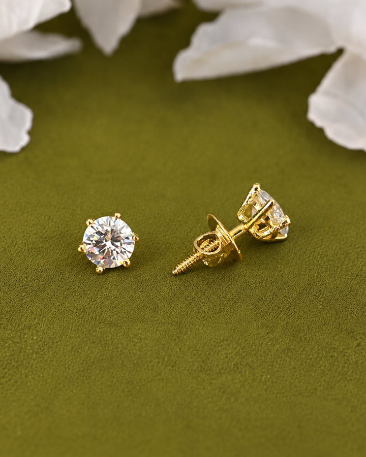 A pair of gold stud earrings with round clear gemstones, one shown from the front and the other from the side/back. They are placed on a textured olive green surface with blurred white flower petals in the background.