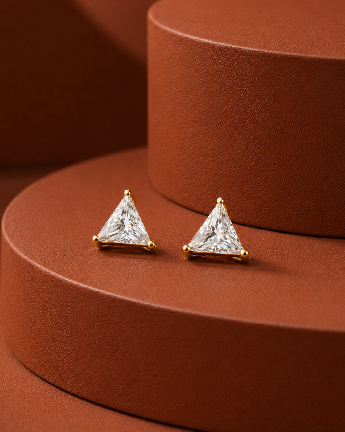 A pair of gold triangle stud earrings with clear triangular gemstones, displayed on a textured terracotta-colored circular pedestal.