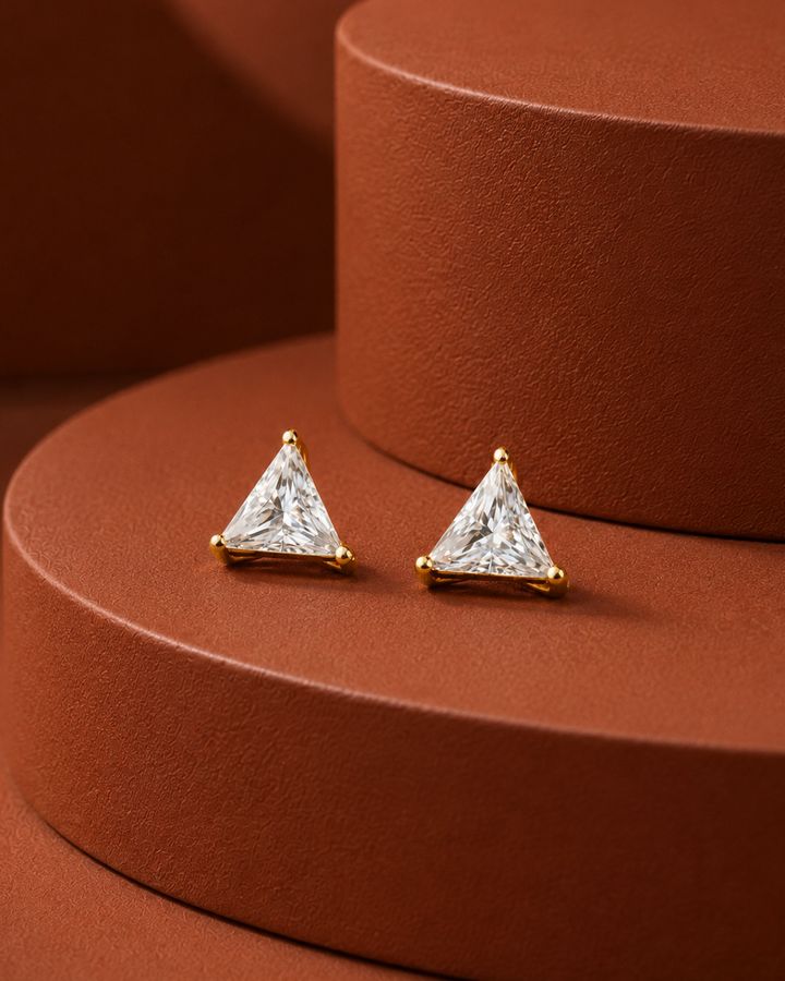 A pair of gold triangle stud earrings with clear triangular gemstones, displayed on a textured terracotta-colored circular pedestal.