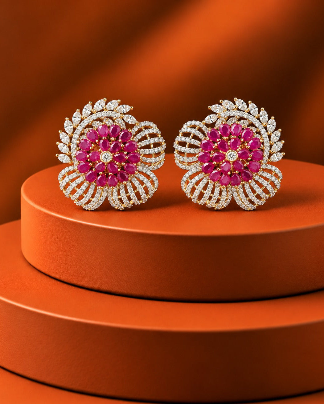 A pair of ornate floral stud earrings with bright pink gemstone centers and silver-toned pavé settings, displayed on stacked orange cylindrical pedestals against a warm orange background.