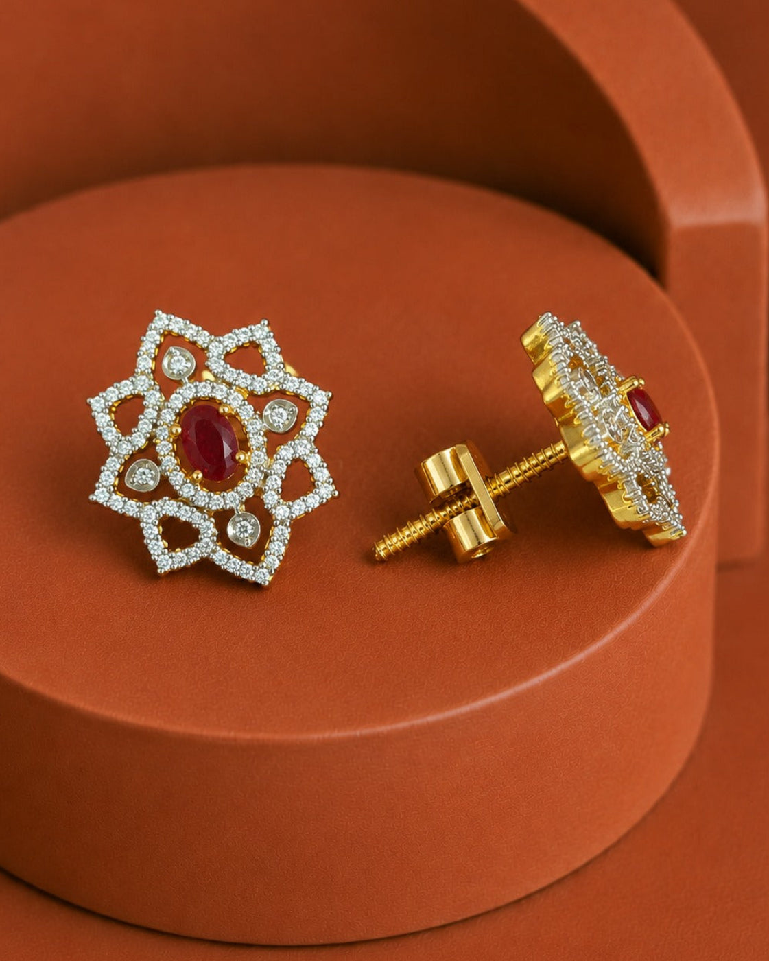 A pair of ornate gold and white stone stud earrings with a central red oval gemstone, displayed on a terracotta-colored geometric pedestal. One earring is shown from the front, and the other is turned to show the gold post and backing.