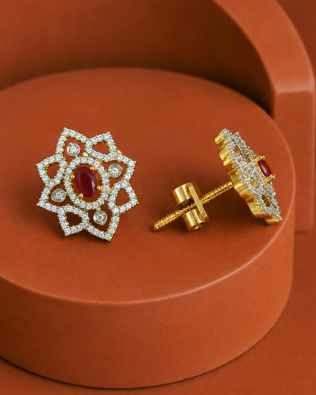 A pair of ornate gold and white stone stud earrings with a central red oval gemstone, displayed on a terracotta-colored geometric pedestal. One earring is shown from the front, and the other is turned to show the gold post and backing.