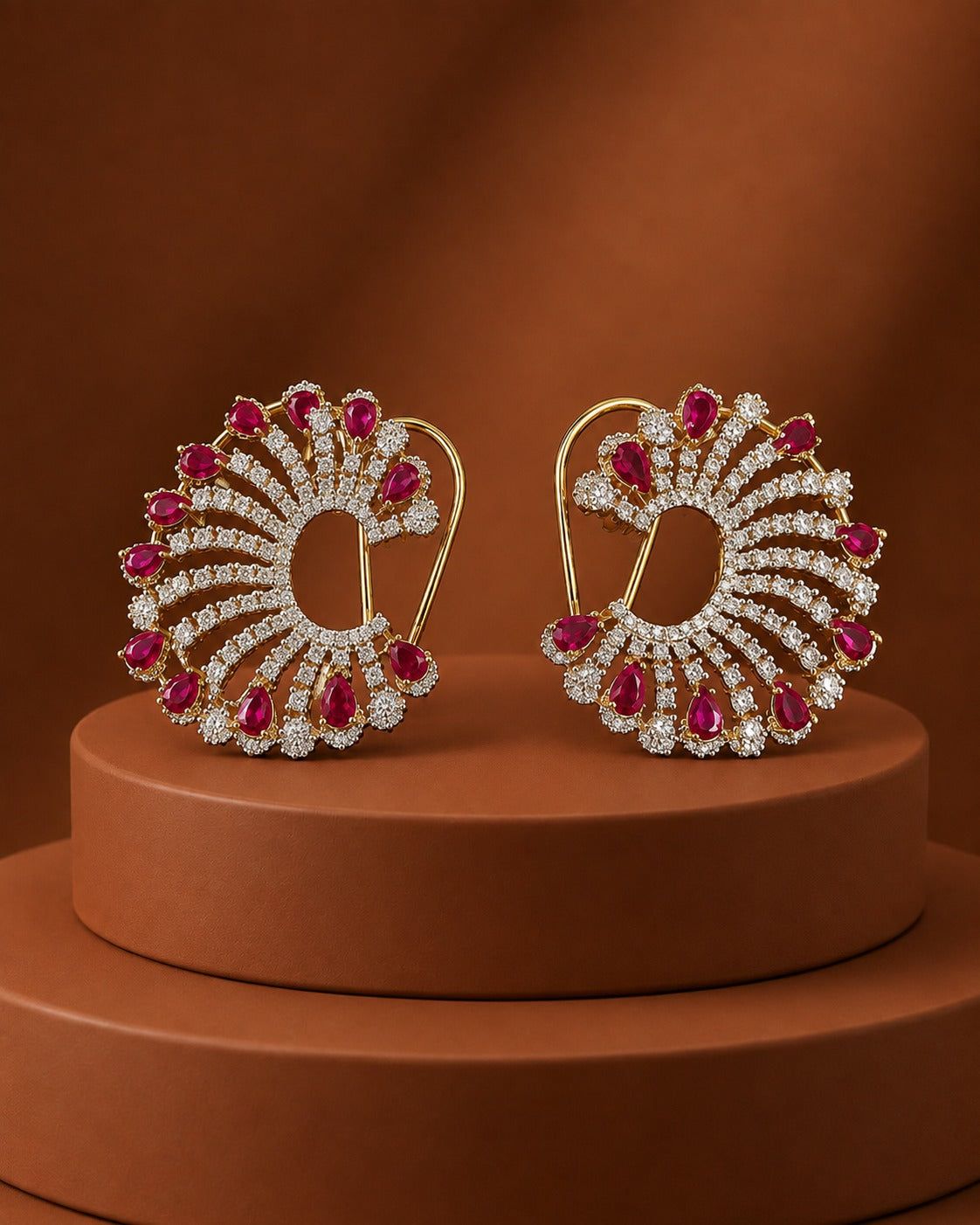 A pair of ornate gold earrings with clear stones and red gemstone accents, displayed on a round brown pedestal against a warm brown background.