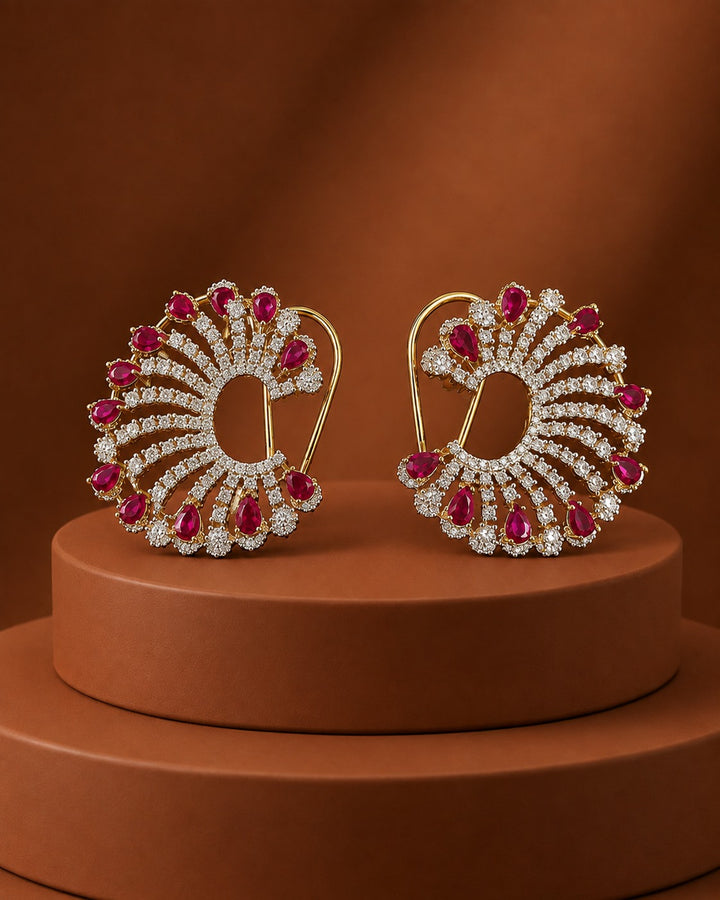 A pair of ornate gold earrings with clear stones and red gemstone accents, displayed on a round brown pedestal against a warm brown background.