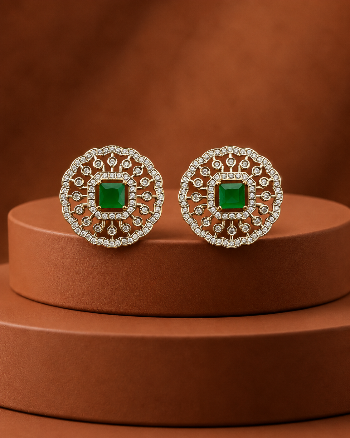 A pair of ornate round stud earrings with square green center stones and intricate silver-toned filigree borders, displayed on a brown cylindrical pedestal against a matching brown background.