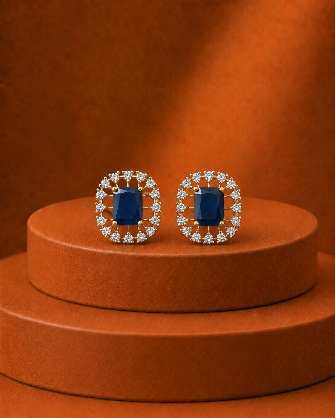 A pair of oval stud earrings with deep blue rectangular center stones surrounded by small clear stones in a gold-tone halo setting, displayed on a round terracotta pedestal against a matching warm brown background.