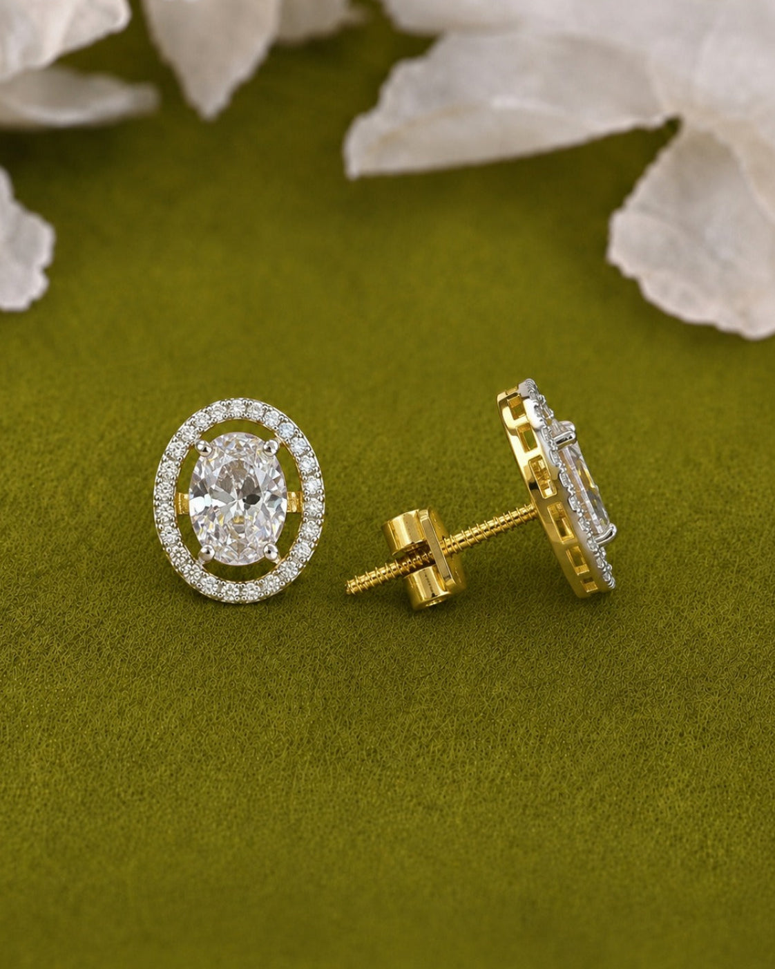 A pair of oval yellow gold stud earrings with a halo of small clear stones and a central oval clear stone, shown on an olive green textured background with white flowers in the background.