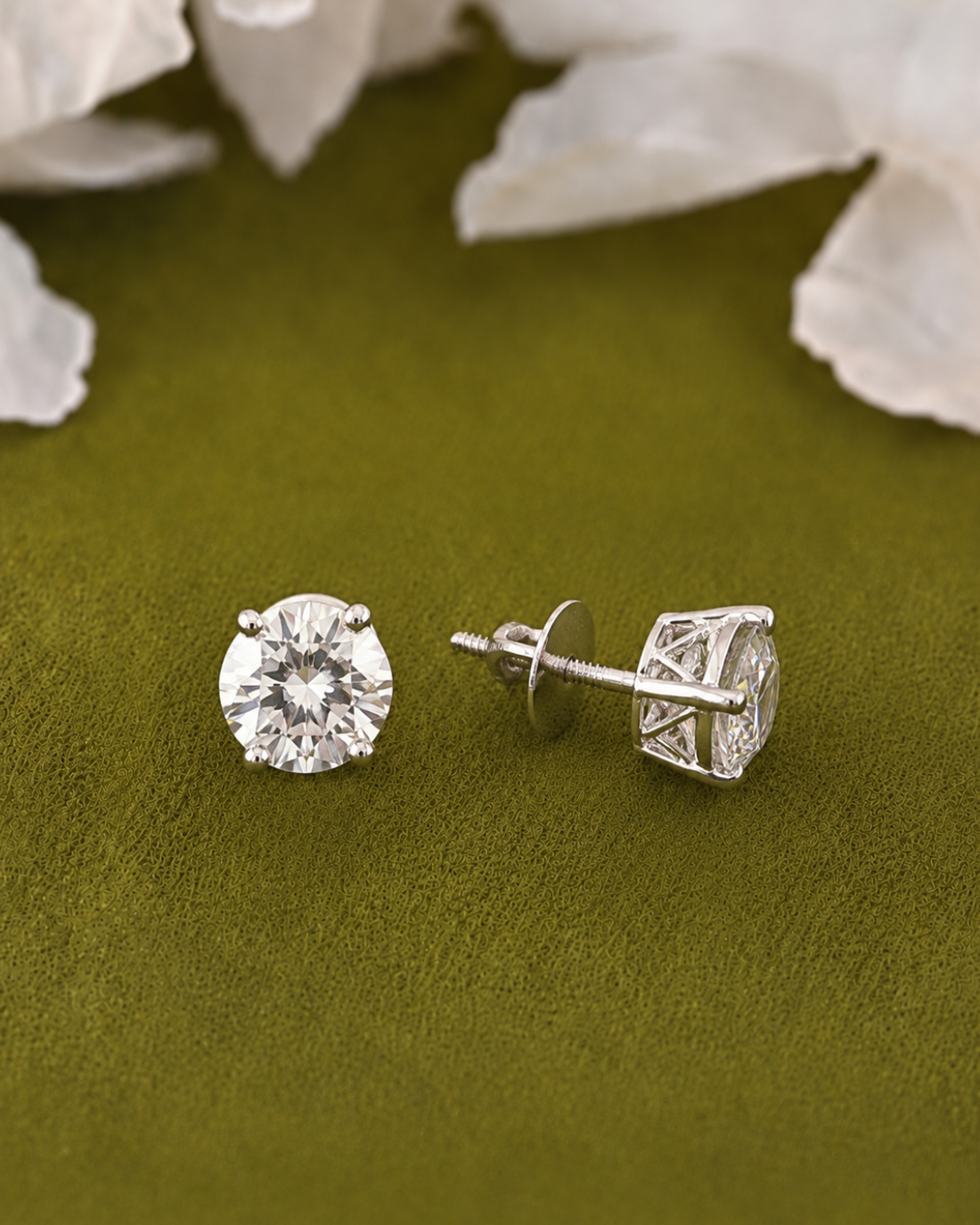A pair of round-cut diamond stud earrings in a silver-toned setting, with one earring facing forward and the other turned to show the back post and butterfly clutch, displayed on a textured olive green surface with soft white flower petals in the background.