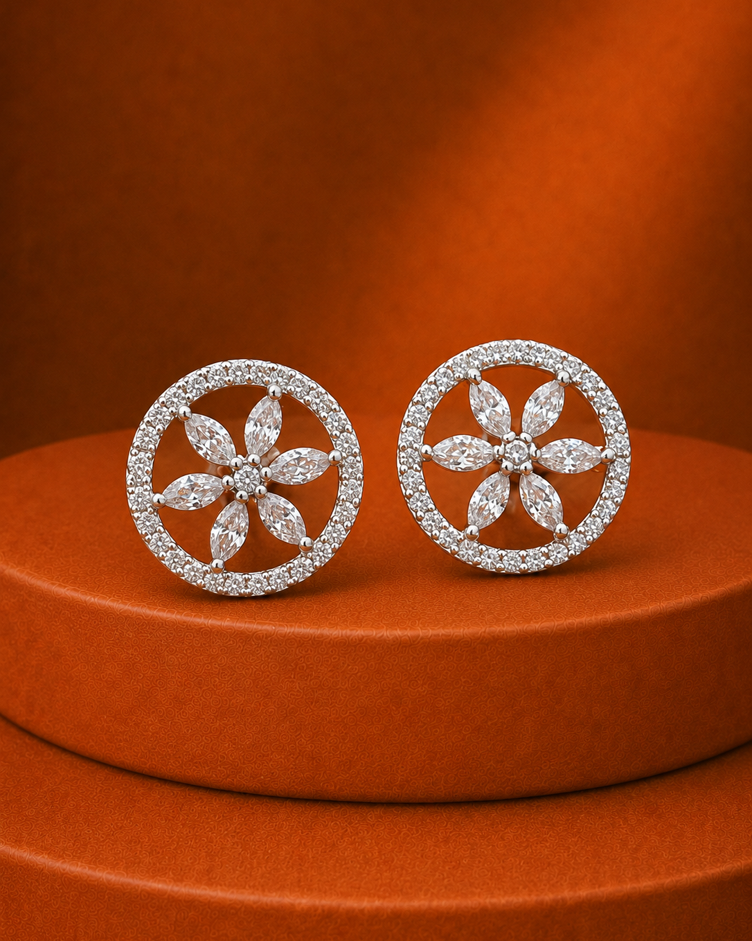 A pair of round floral stud earrings with silver-toned metal and pavé-set clear stones, displayed on a round orange pedestal against a matching orange background.