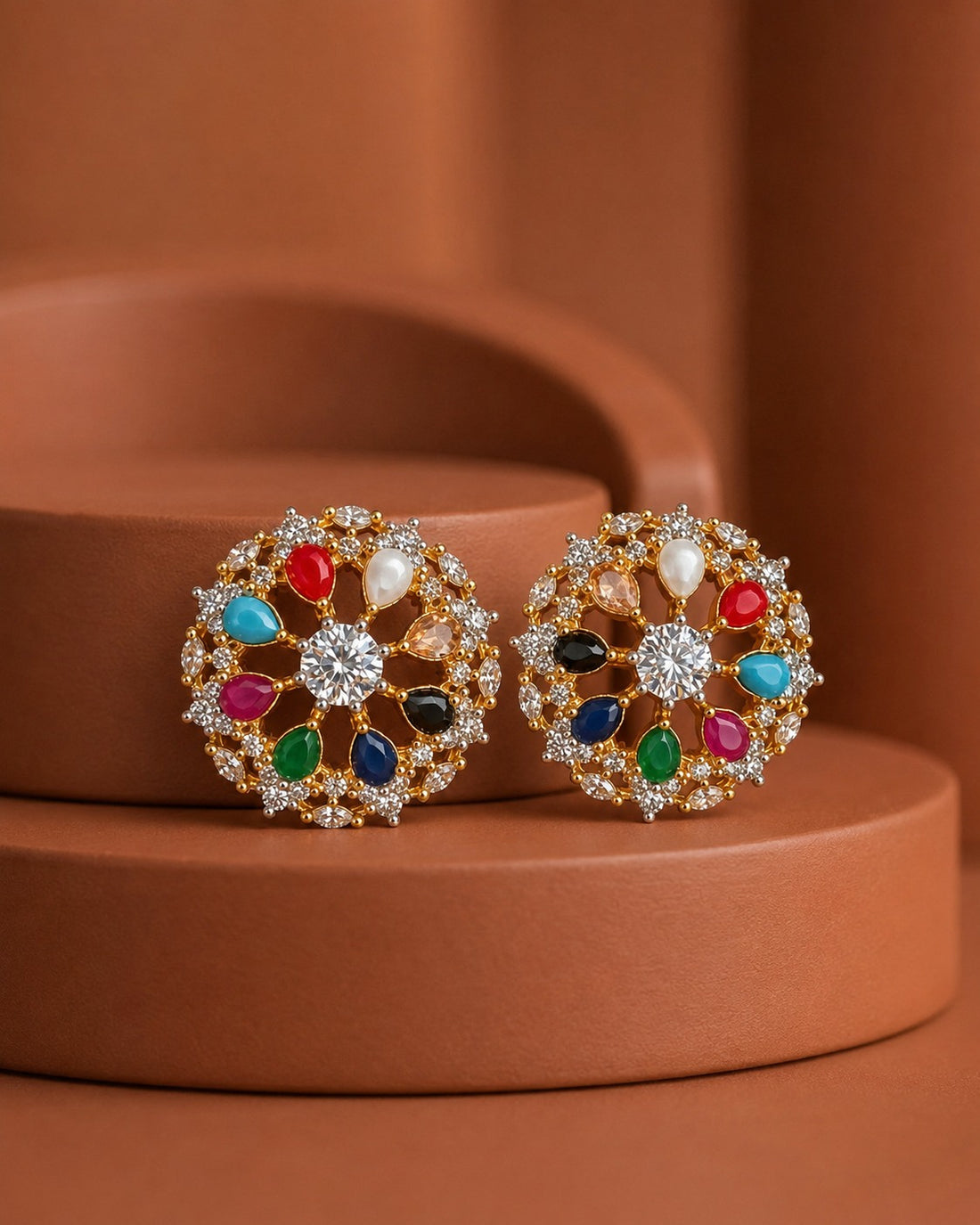 A pair of round gold-toned stud earrings decorated with colorful gemstones and clear crystals, displayed on a terracotta-colored tiered pedestal.