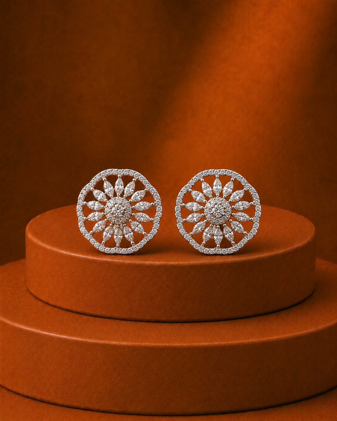 A pair of round silver-toned earrings paved with clear stones in a sunburst pattern, displayed on a round brown pedestal against a matching brown background.