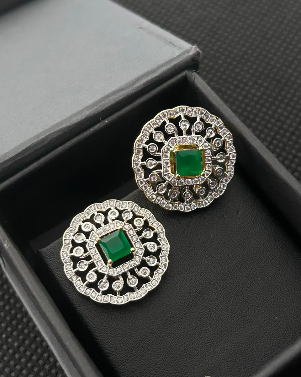 A pair of round silver-toned earrings with green square-cut center stones and surrounding clear stones, displayed in a black jewelry box.