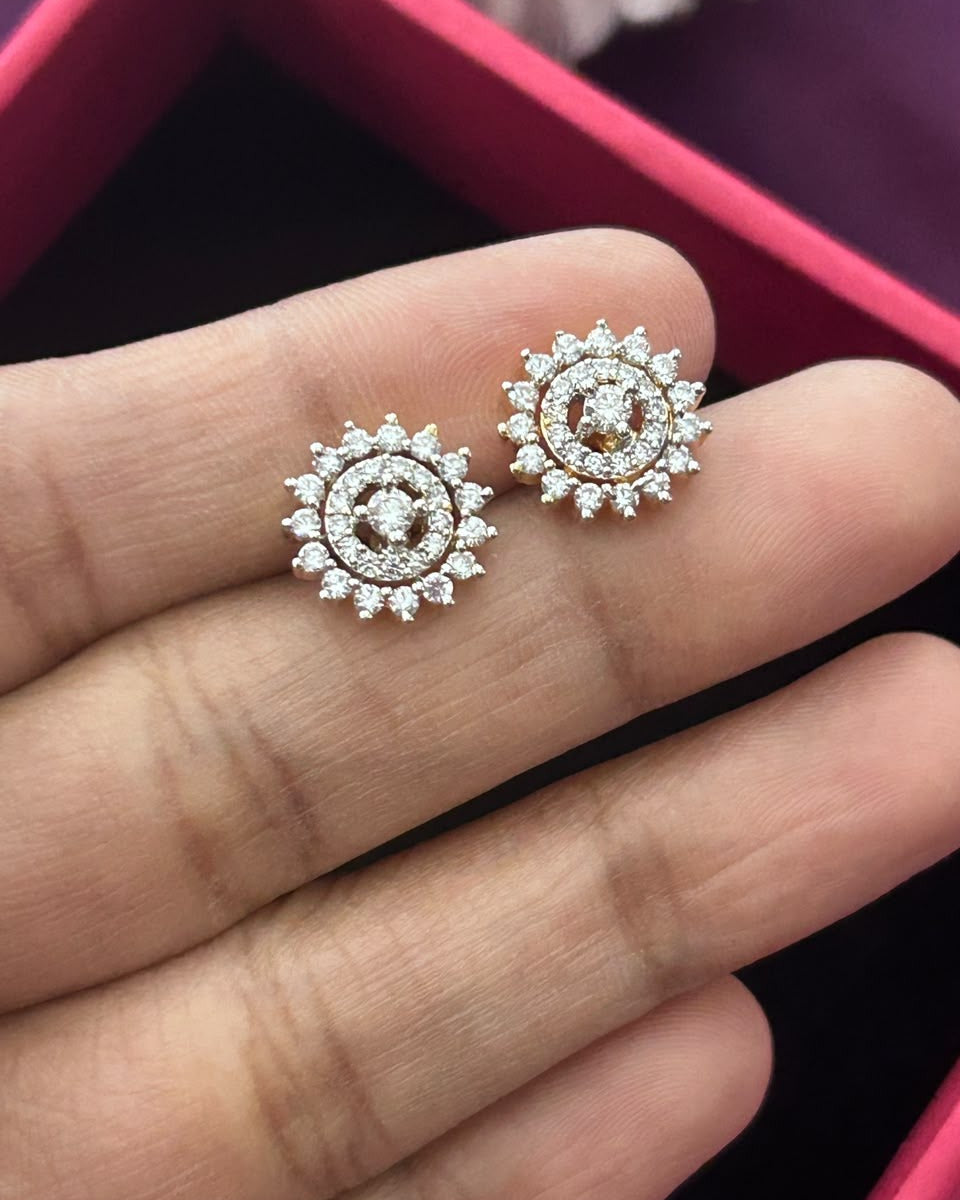A pair of round stud earrings with a silver-tone floral or sunburst design set with small clear stones, held on a person's fingers over a pink jewelry box.