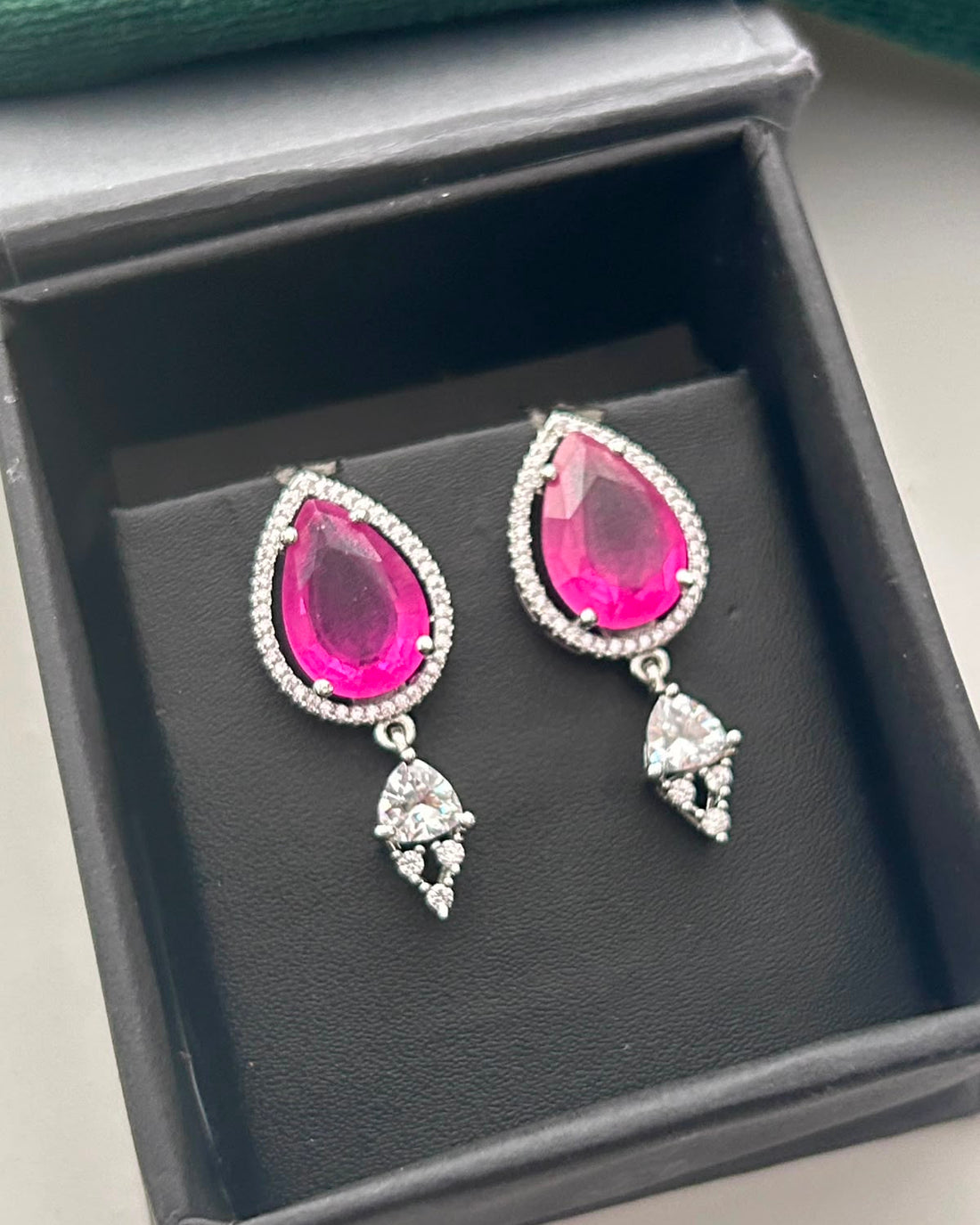 A pair of silver-tone teardrop dangle earrings with vibrant pink gemstones and small clear stones, displayed inside a black jewelry box.