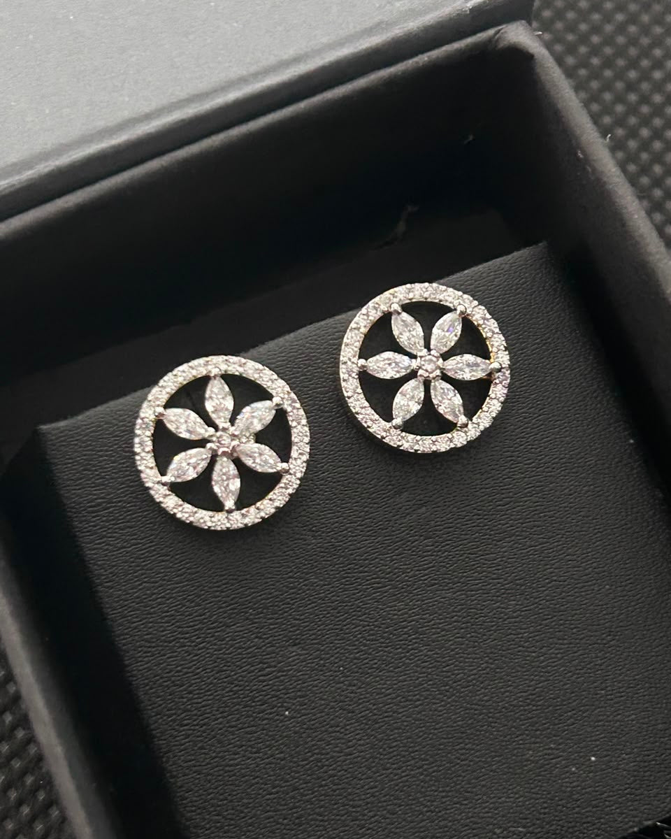 A pair of silver-tone circular stud earrings with a floral six-petal design, set with small clear stones, displayed on a black textured jewelry box.