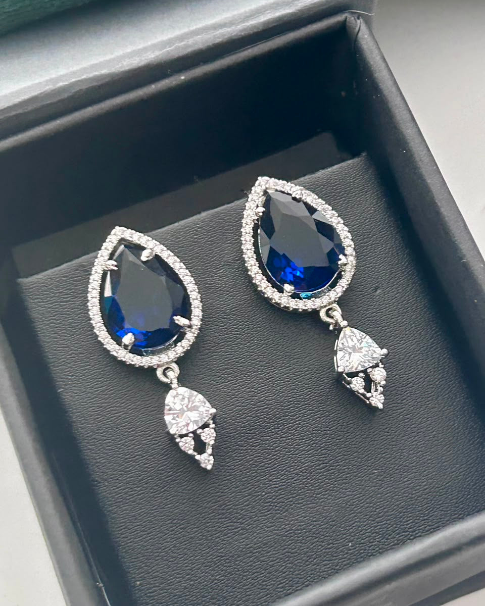 A pair of silver-tone teardrop dangle earrings with deep blue teardrop gemstones and clear pavé accents, displayed inside a black jewelry box.
