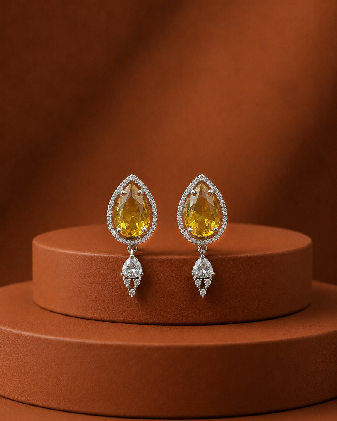 A pair of silver-tone teardrop earrings with large yellow gemstones and small clear stones, displayed on a round terracotta pedestal against a matching warm brown background.