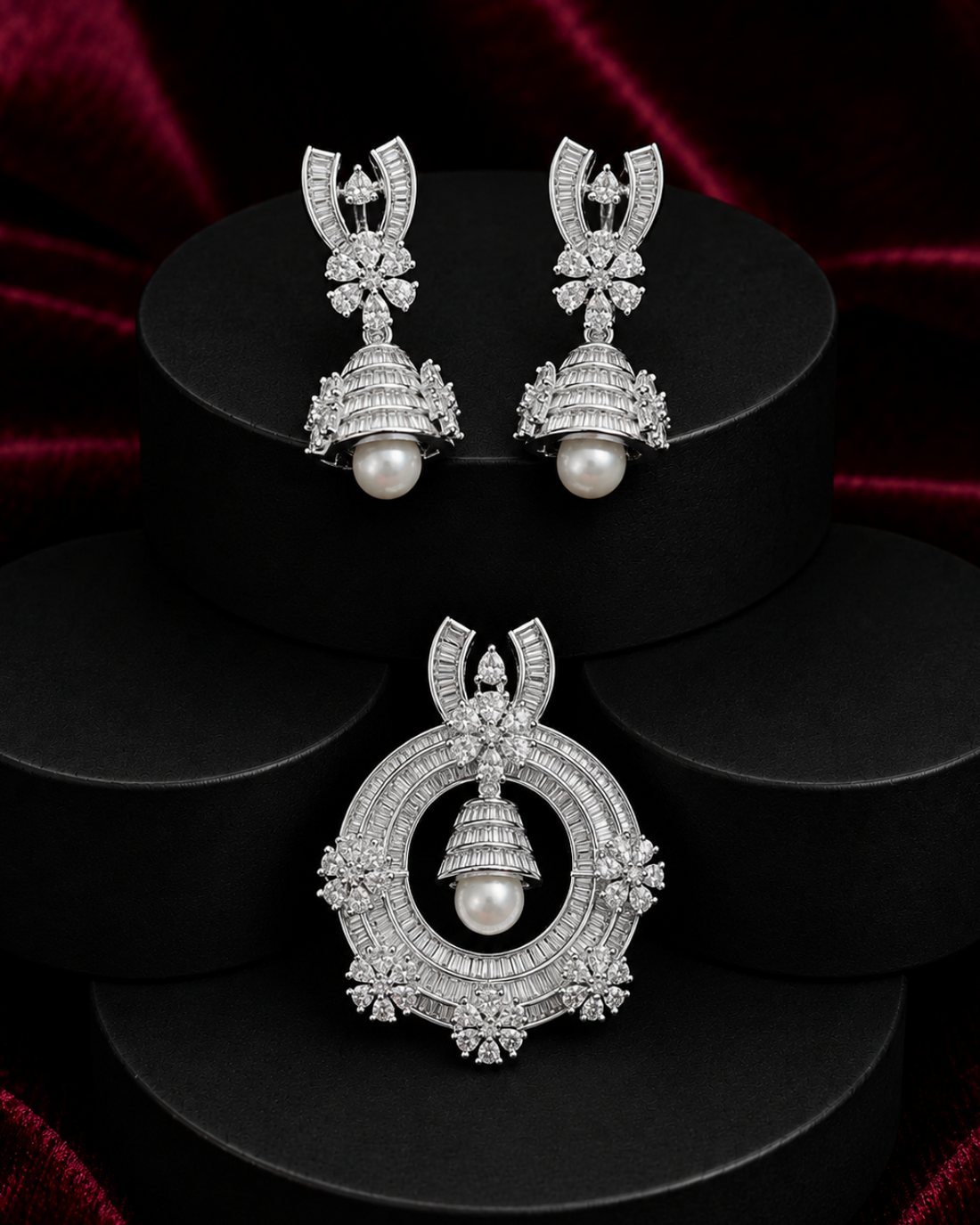 A pair of silver-toned Jhumka earrings and a matching Jhumka pendant set with clear stones and white pearls, displayed on black cylindrical pedestals against a dark red velvet background.