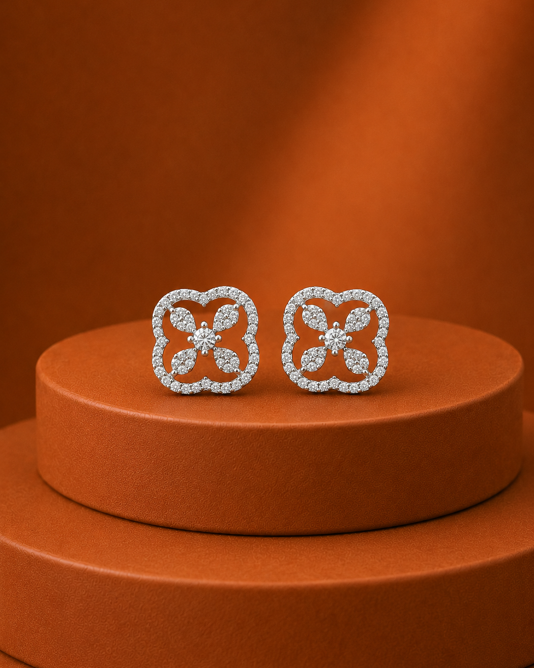 A pair of silver-toned clover-shaped stud earrings set with clear stones, displayed on a round terracotta-colored pedestal against a matching warm brown background.