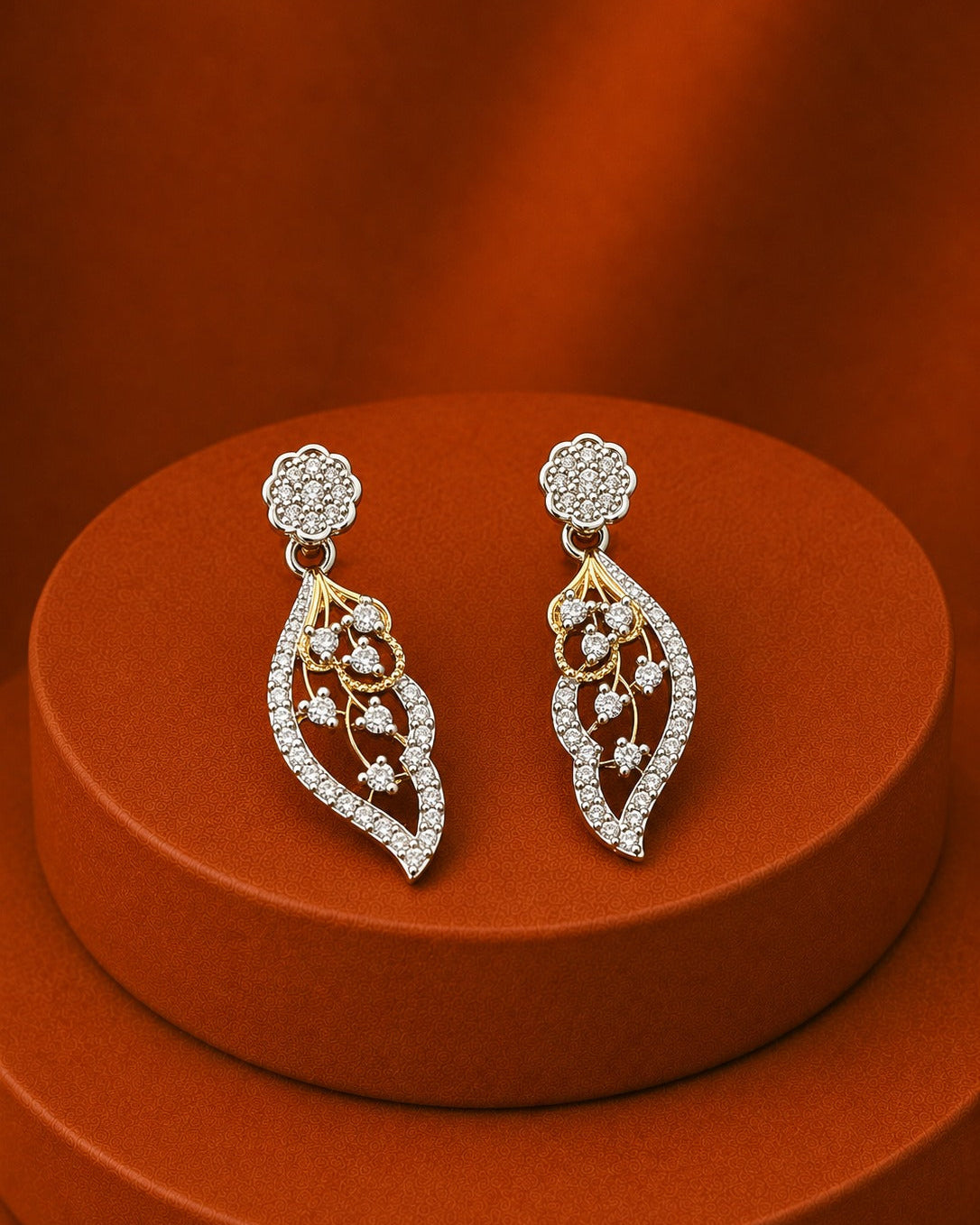 A pair of silver-toned dangle earrings with gold-toned accents and clear pavé stones, displayed on a round orange-brown pedestal against a matching warm brown background.