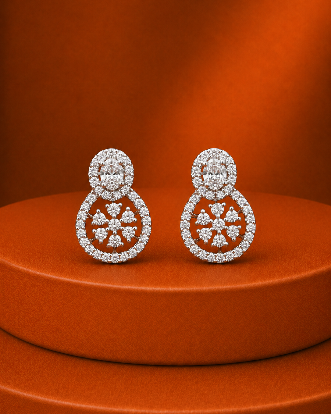 A pair of silver-toned double-halo stud earrings paved with clear stones, featuring a circular halo at the top and a floral-like pattern at the bottom, displayed on a textured orange pedestal against a matching orange background.