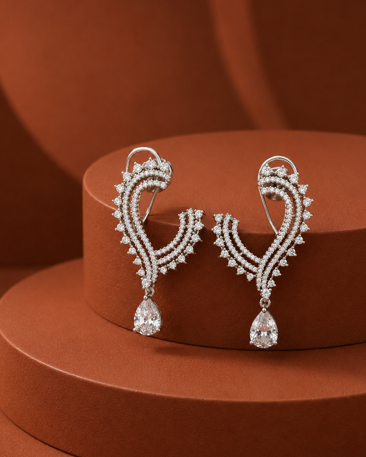A pair of silver-toned drop earrings with curved, pavé-set clear stones and teardrop-shaped dangling drops, displayed on a terracotta-colored circular pedestal.