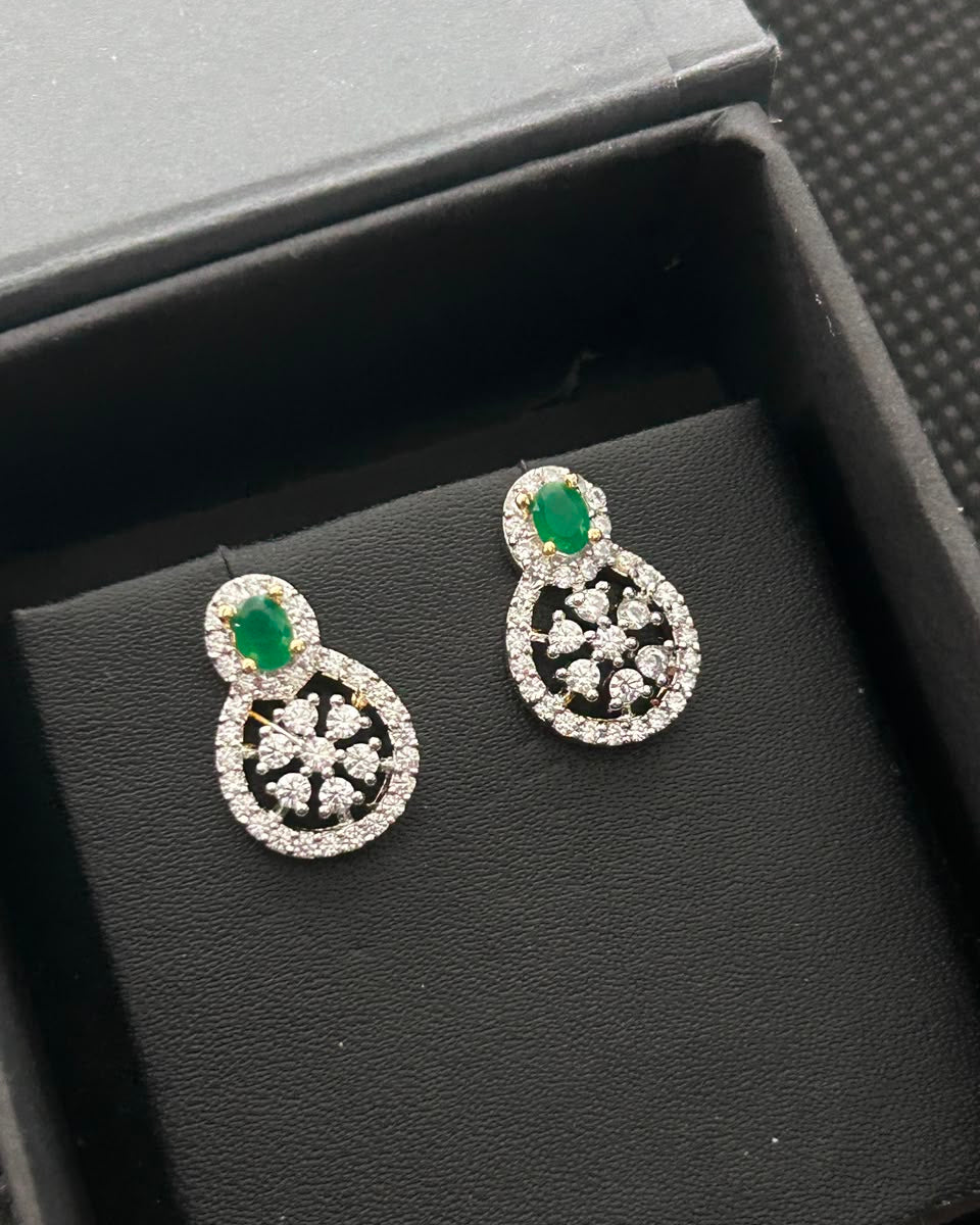 A pair of silver-toned drop earrings with oval-cut green stones and pavé-set clear stones, displayed on a black textured jewelry box.