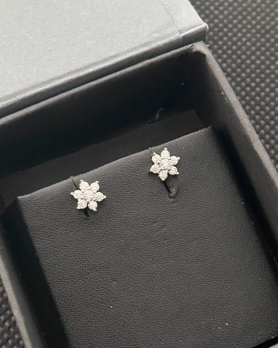 A pair of silver-toned flower-shaped stud earrings with clear stones, displayed on a black textured jewelry box insert.