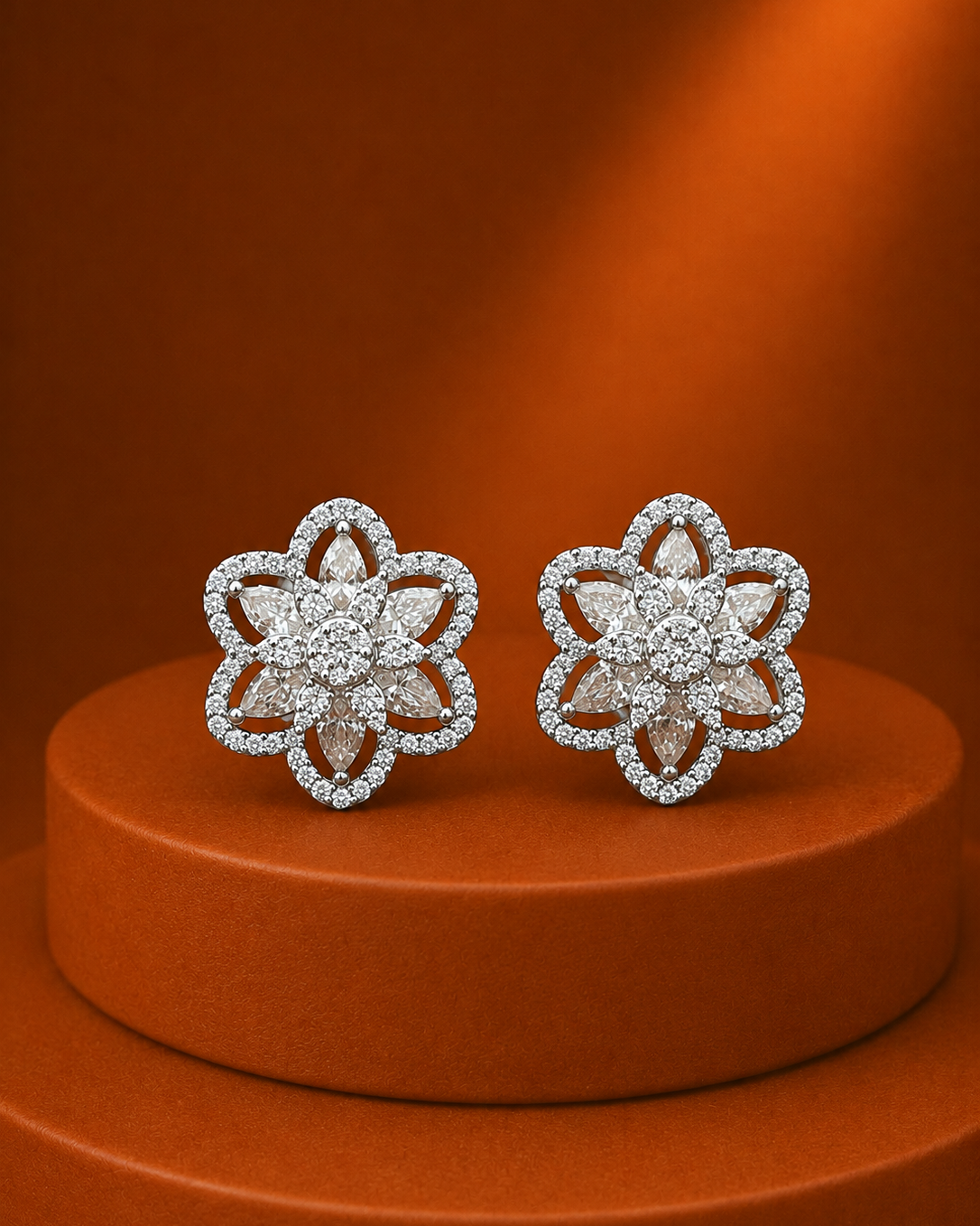 A pair of silver-toned flower-shaped stud earrings paved with clear stones, displayed on a round orange pedestal against a matching orange background.