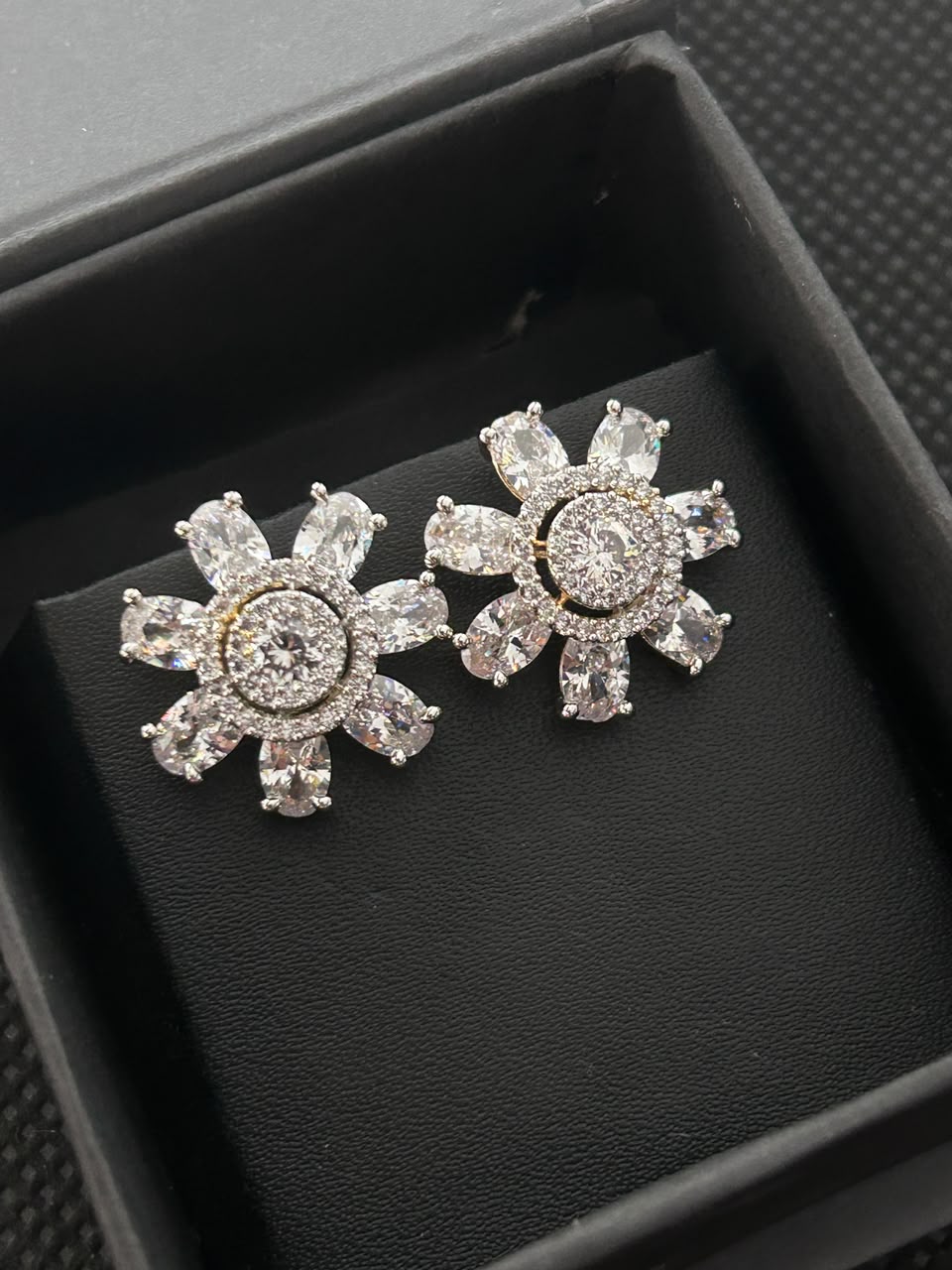 A pair of silver-toned flower-shaped stud earrings with clear crystals, displayed on a black velvet insert inside a black jewelry box.