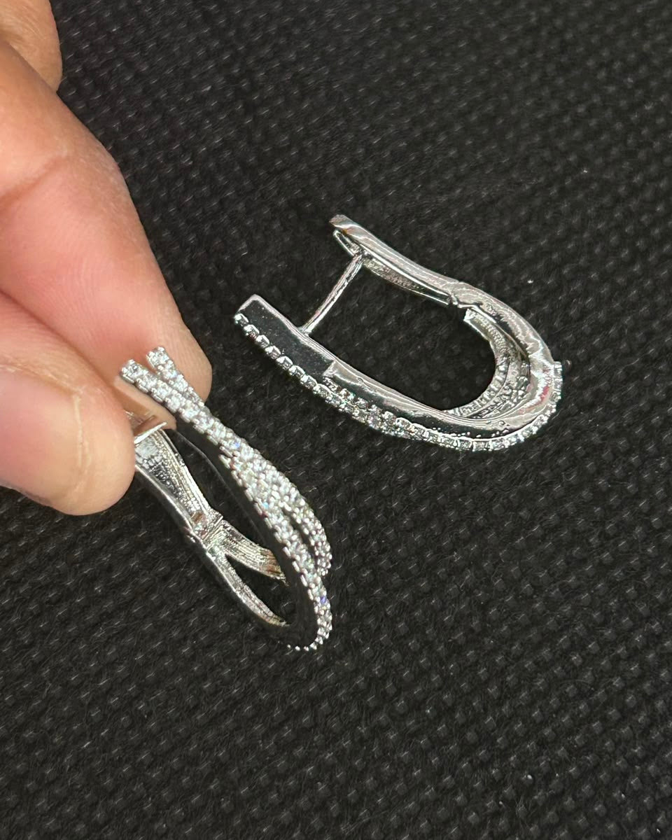 A pair of silver-toned hoop earrings with pavé-set clear stones, shown with one earring held between fingers against a textured black background.