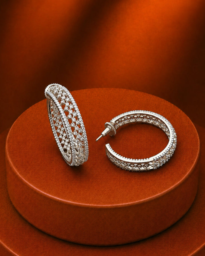 A pair of silver-toned hoop earrings covered in small clear stones, displayed on a round orange-brown pedestal against a matching draped background.