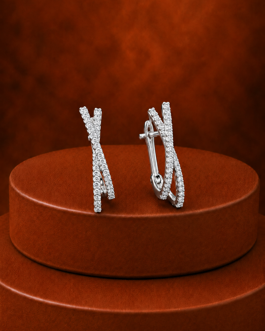 A pair of silver-toned hoop earrings with multiple rows of small clear stones arranged in an abstract curved pattern, displayed on a round brown pedestal against a textured brown background.