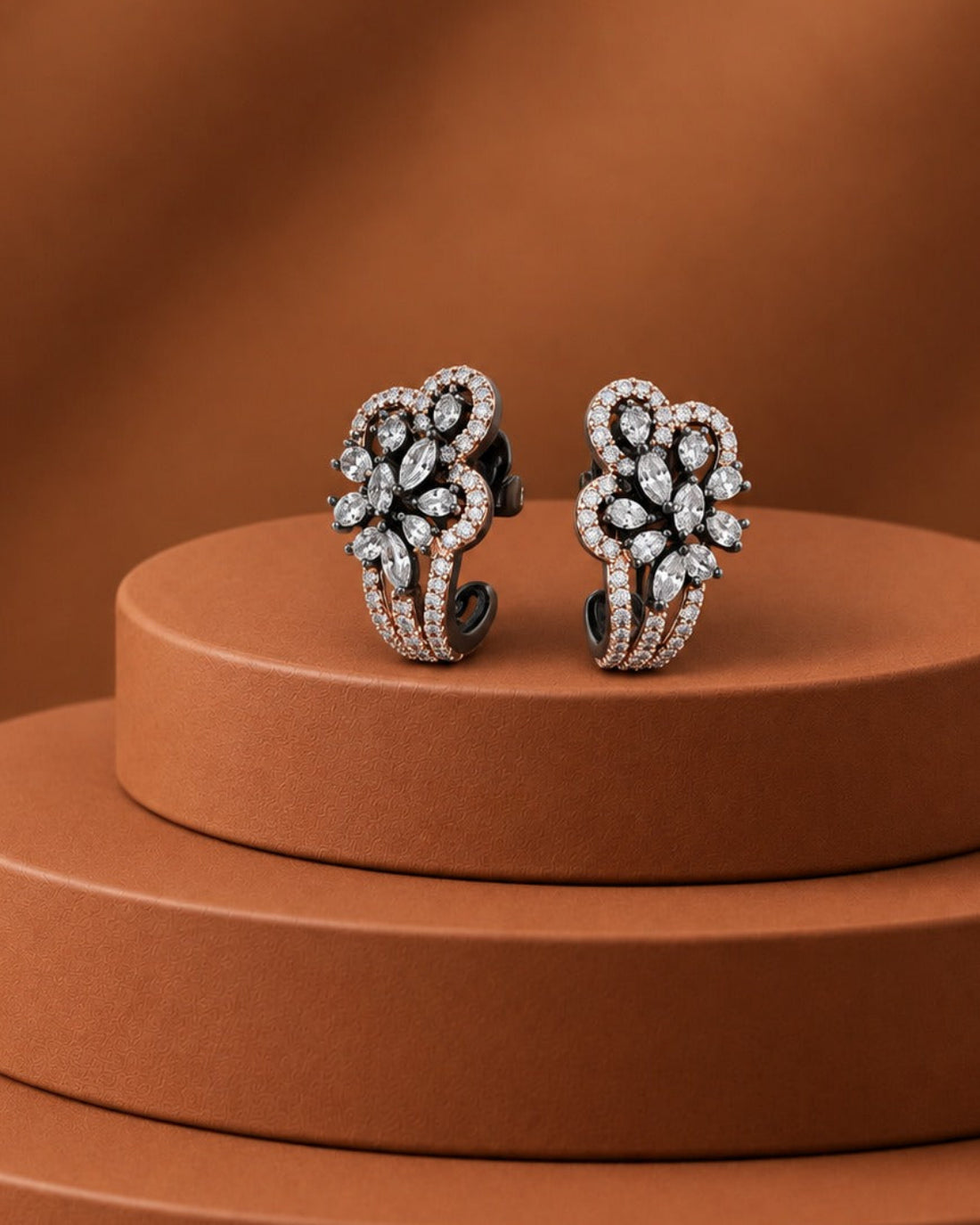 A pair of silver-toned huggie hoop earrings adorned with pavé-set clear stones, displayed on a tiered brown pedestal against a matching brown background.