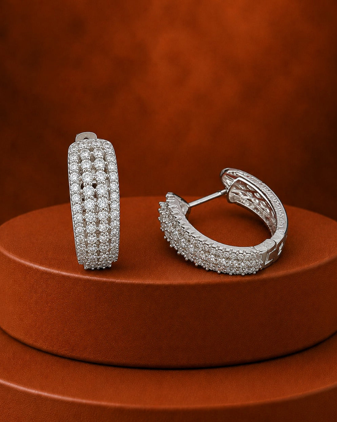 A pair of silver-toned huggie hoop earrings paved with clear stones, displayed on a round terracotta pedestal against a textured orange-brown background.