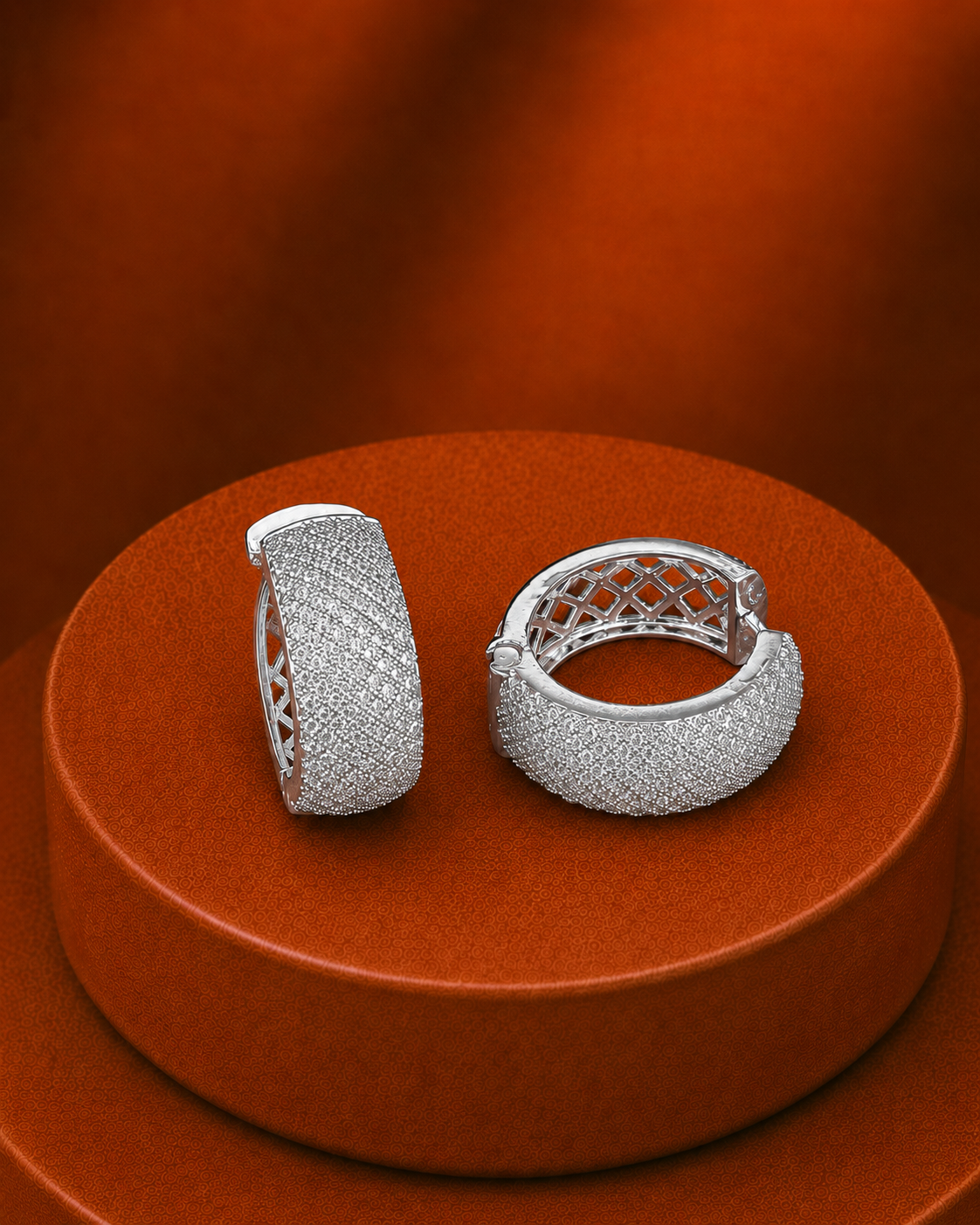 A pair of silver-toned pavé hoop earrings displayed on a round orange pedestal against a matching orange background.