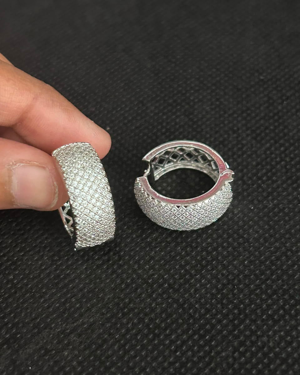 A pair of silver-toned pavé huggie hoop earrings, with one earring held by a hand and the other resting on a textured black background.