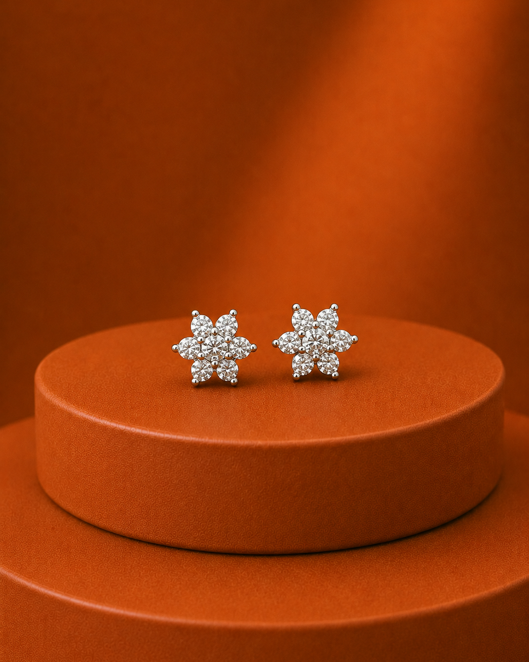 A pair of silver-toned snowflake stud earrings set with clear crystals, displayed on a round orange pedestal against a matching orange background.