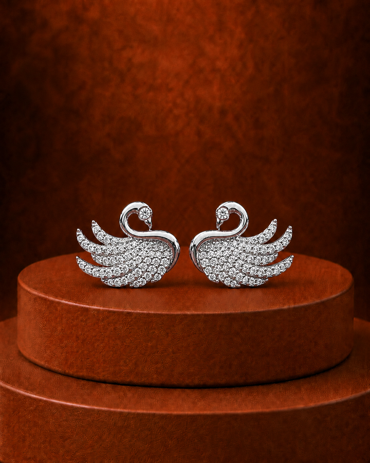 A pair of silver-toned swan-shaped earrings paved with small clear stones, displayed on a round brown pedestal against a textured brown background.