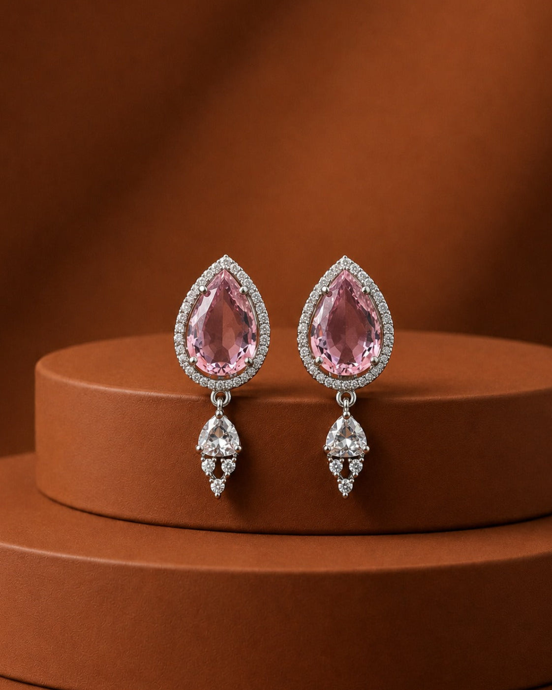 A pair of silver-toned teardrop dangle earrings with pink gemstones and clear accent stones, displayed on a round brown pedestal against a warm brown background.