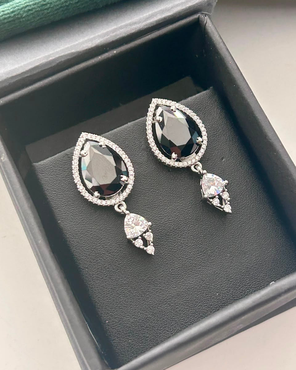 A pair of silver drop earrings with teardrop-shaped black gemstones surrounded by small clear stones, displayed in a black jewelry box.