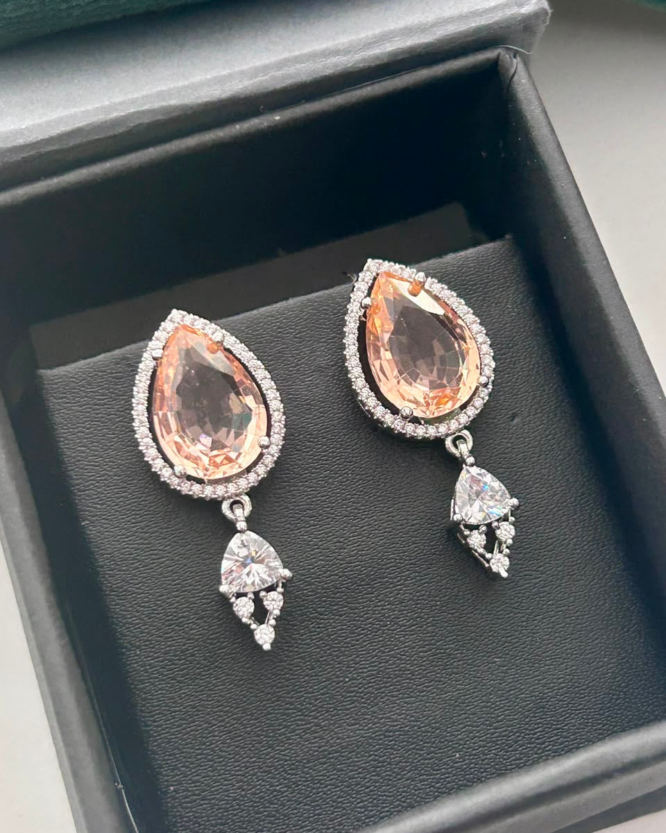 A pair of silver drop earrings with teardrop-shaped peach-pink gemstones surrounded by small clear stones, displayed on a black jewelry box.