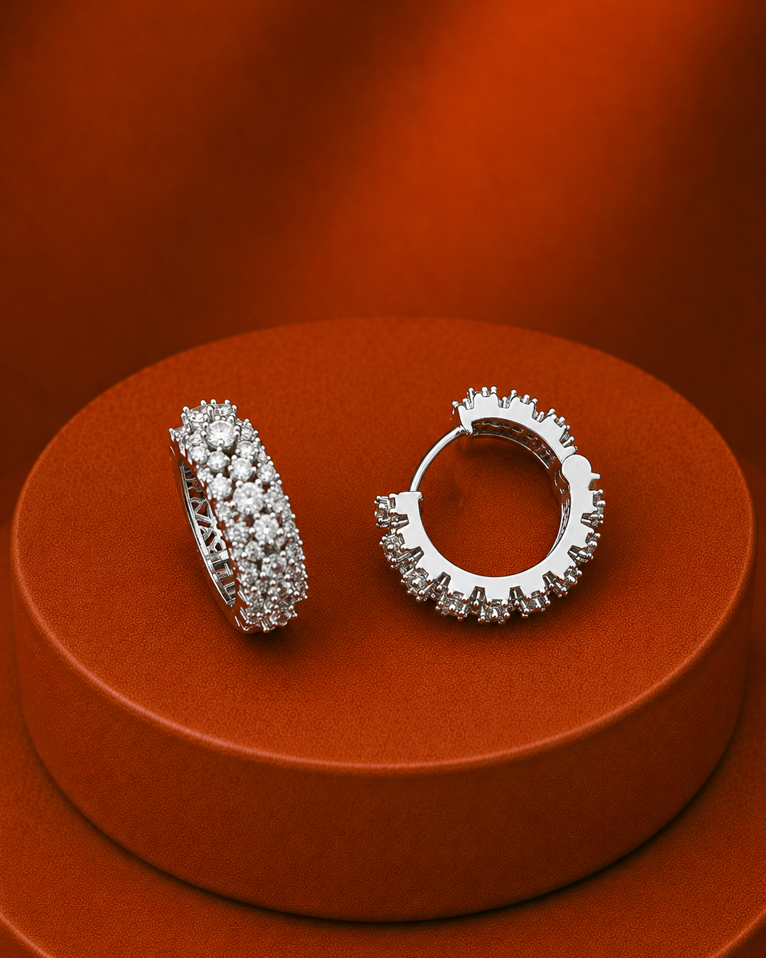 A pair of silver hoop earrings covered in small clear stones, displayed on a round orange pedestal against a matching orange background.