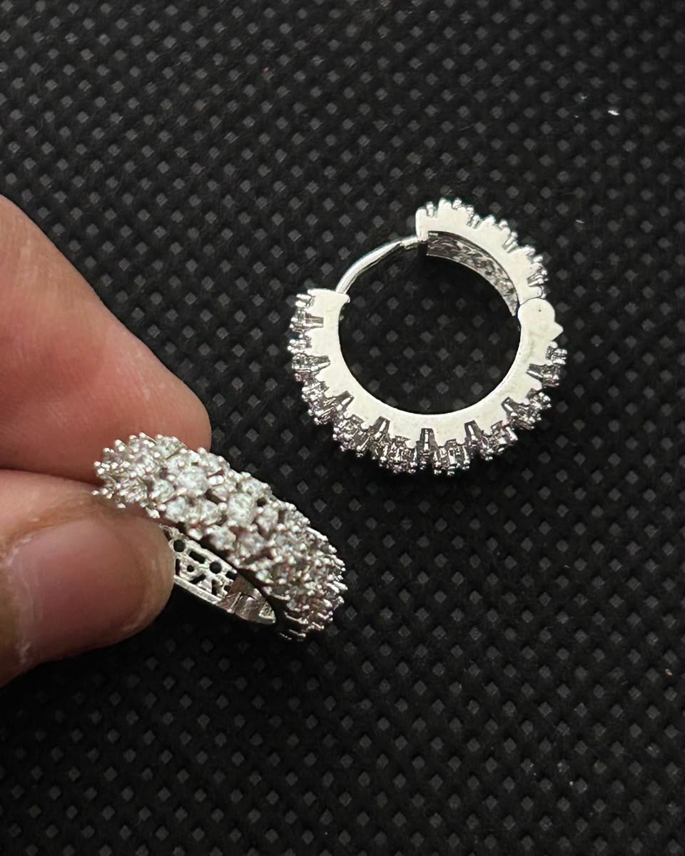 A pair of silver hoop earrings paved with clear stones, with one earring displayed on a textured black background and the other held by a person's fingers to show scale.