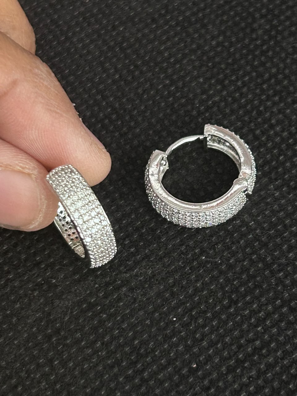 A pair of silver huggie hoop earrings covered in small clear stones, with one earring held between fingers and the other resting on a black textured surface.